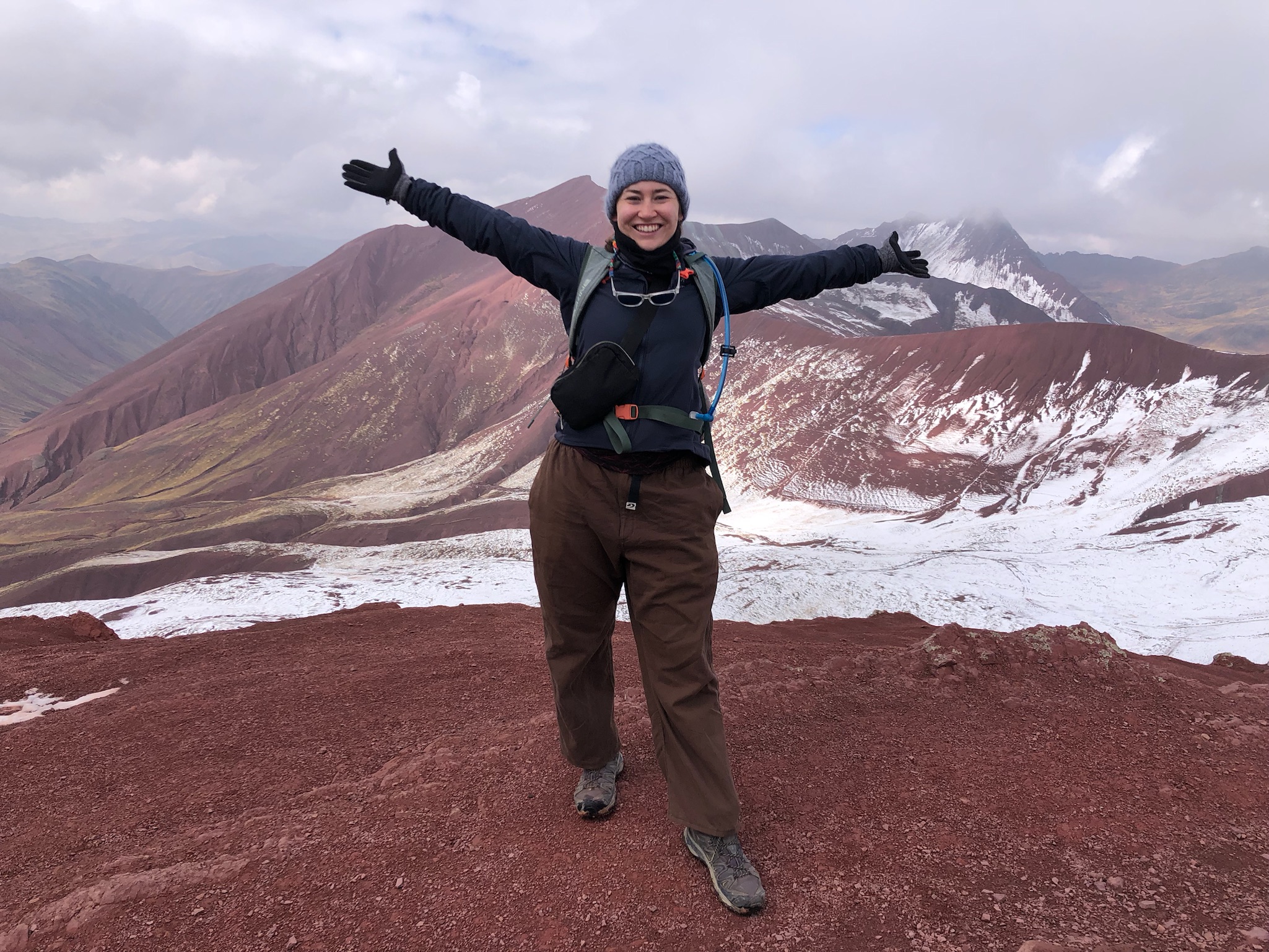 Leah Huey smiling with arms outstretched in front of a turquoise alpine lake and snow-capped peaks
