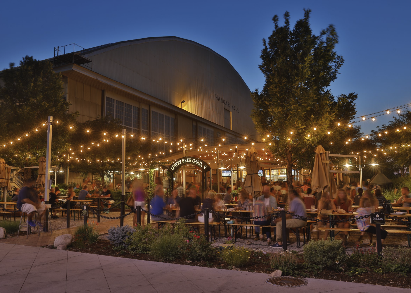 Lowry Beer Garden at dusk, with string lights glowing over crowded picnic tables in front of Hangar No. 2